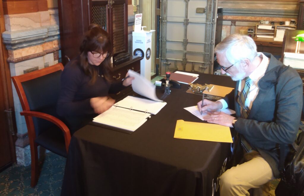 Tom O'Donnell kneels to sign paperwork at the Iowa Secretary of State's office, while an office worker counts his petition pages.
